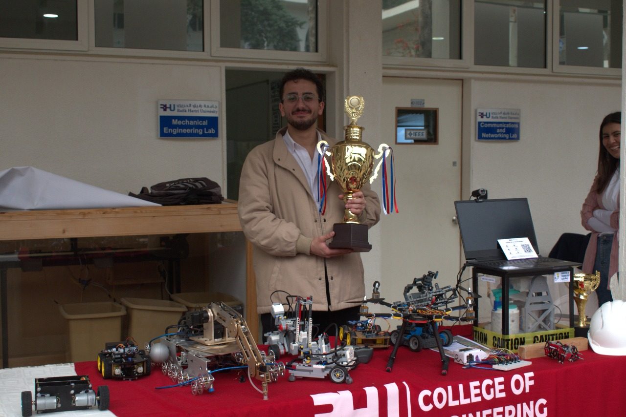 Rami Kronbi holding a large trophy in the RHU mechanical engineering lab, in front of a table of competition robots.