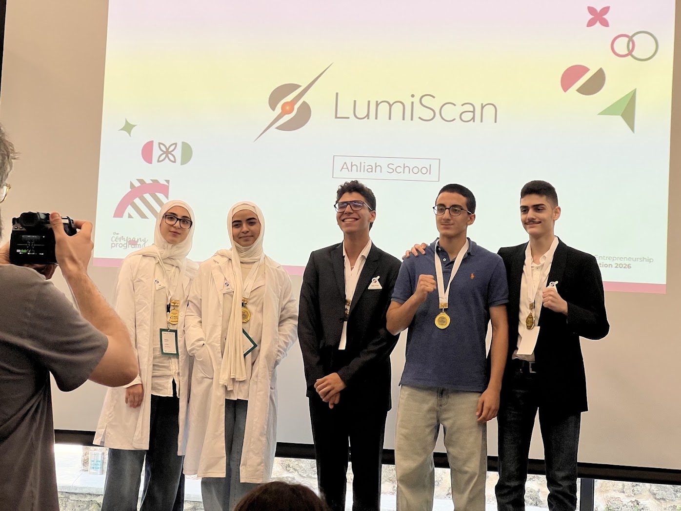 The LumiScan team from Ahliah School on stage at the INJAZ Lebanon final, wearing medals, in front of a screen showing their logo.